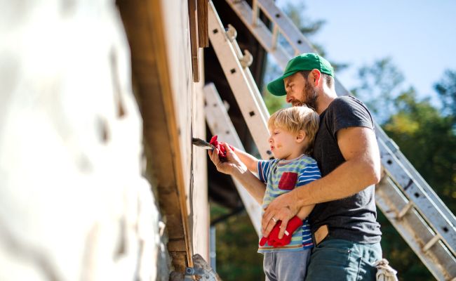 A side view of father and toddler boy outdoors in summer, painting wooden house.