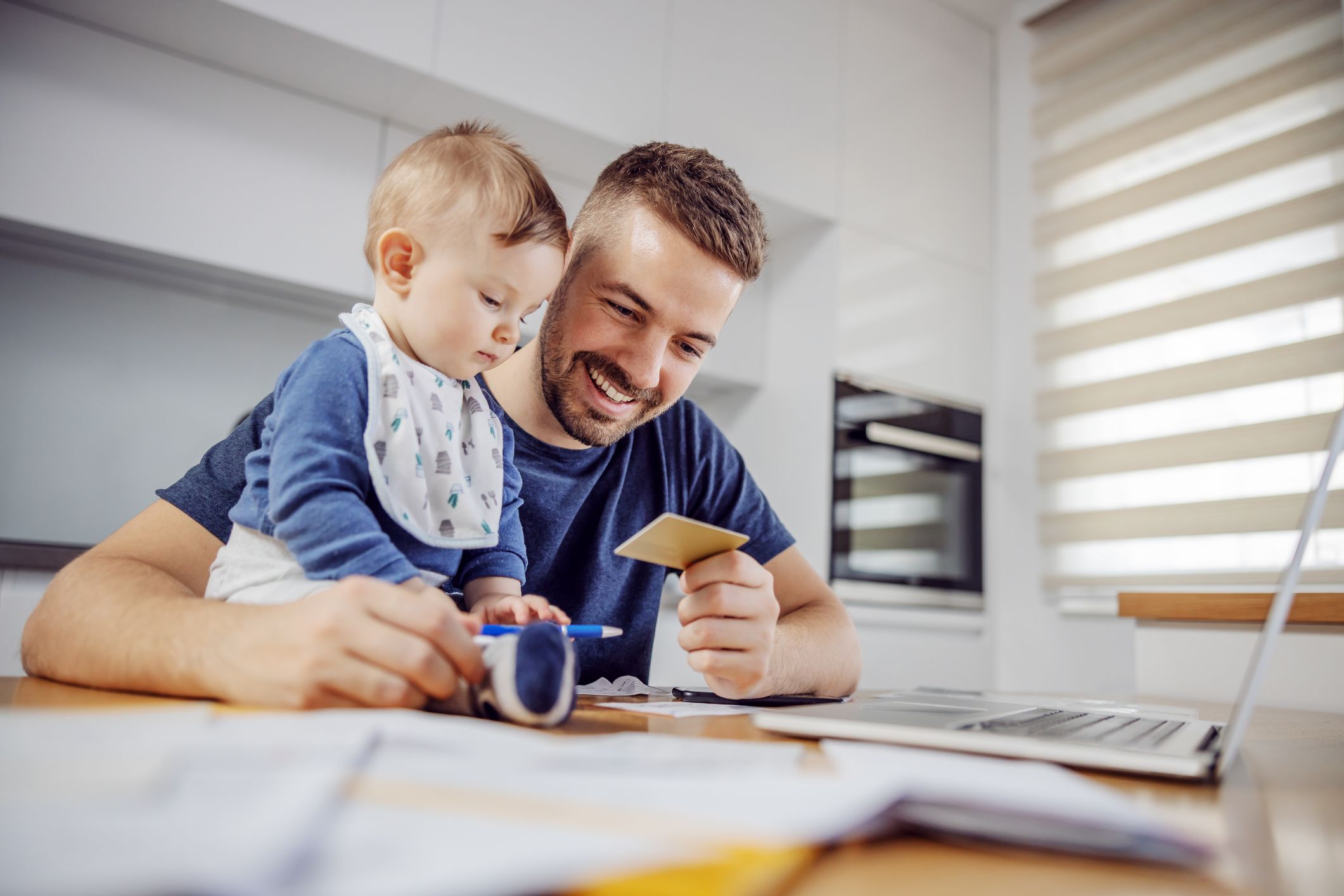 Young attractive bearded father sitting at dinning table with his beloved adorable son and paying bills online. His boy holding pen and trying to help him.