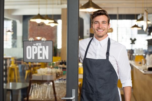 Portrait of smiling owner standing at his restaurant gate with open signboard. Young entrepreneur leaning at the cafeteria door and looking at camera. Chef or waiter standing in front of coffee shop.